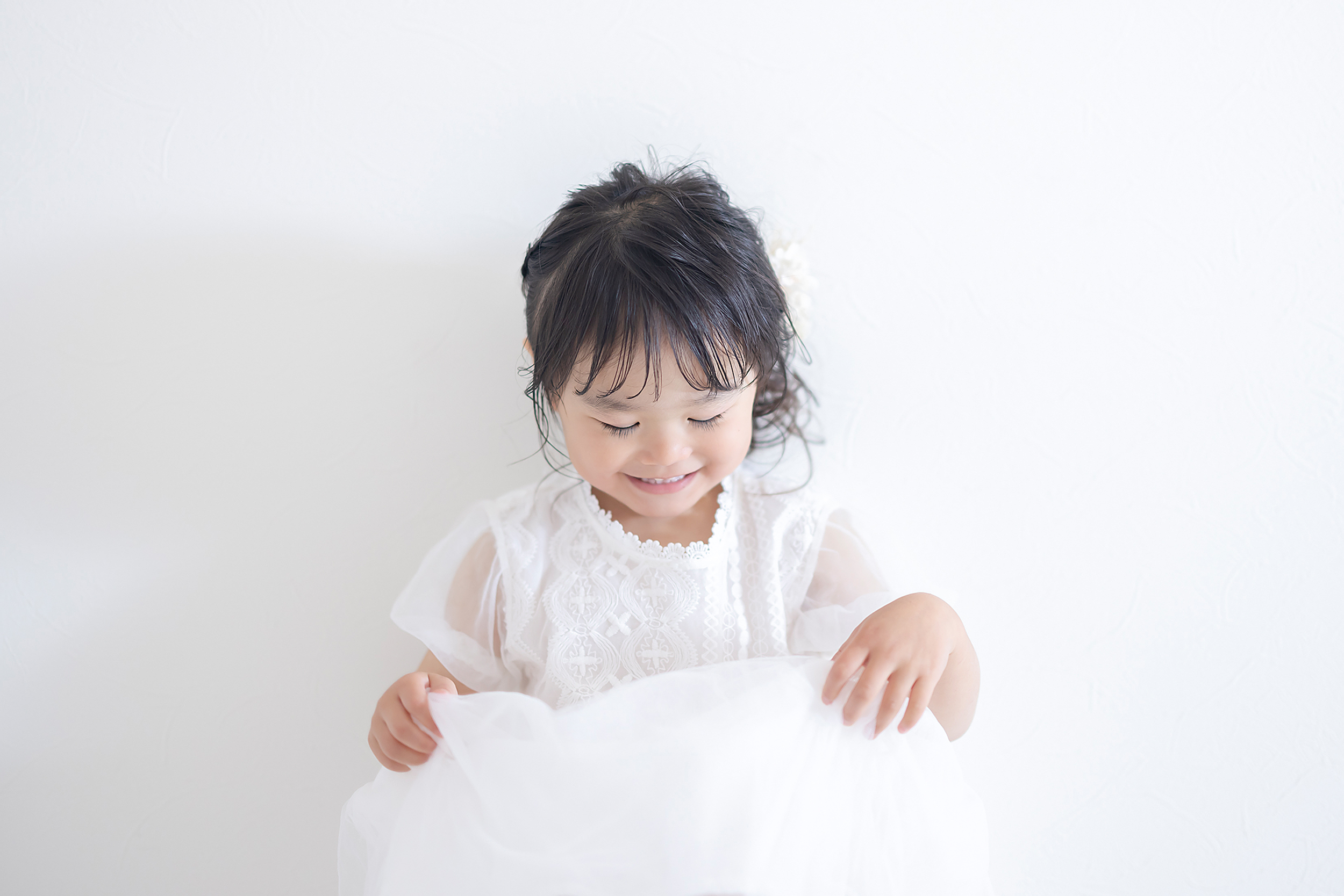 白いドレスを着て笑顔を見せる3歳の女の子。自然光がやさしく包むスタジオ撮影。